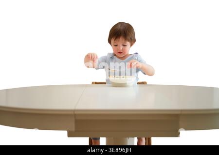 Kleinkind Junge, der gesundes Gemüsegericht alleine am weißen Esstisch isst. Kleines Kind, das im Hochstuhl einen Löffel hält. Selbstentwicklung Stockfoto