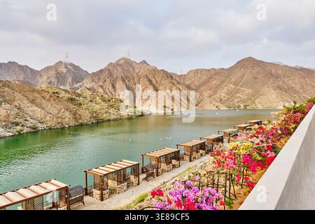 Malerische Raststätte am Al Rafisah Dam mit hölzernen Pavillons, blühenden Blumen und smaragdgrünem Wasser vor dem zerklüfteten Hajar-Gebirge, VAE. Perfektes Konzept Stockfoto
