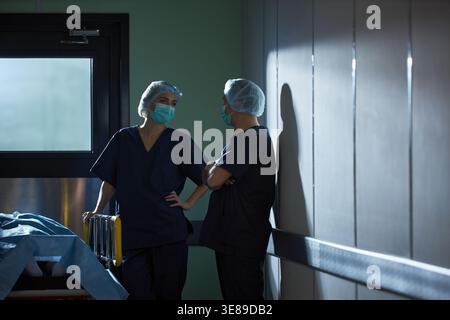 Chirurgeninnen mit Masken und Peelings diskutieren über das Vorgehen neben der Krankenbahre im Korridor. Unterstützung von Teamarbeit im Gesundheitswesen, Personal, Chirurgie, Notfallmarketing Stockfoto