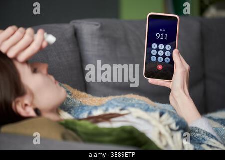 Junge Erwachsene kaukasische Frau, die auf dem Sofa liegt, das Smartphone hält und 911 anruft, während sie Gewebe greift, Krankheit zeigt oder Notfallreaktion zeigt. Nützlich für Gesundheitswarnungen, Telemedizin, Krisenunterstützung Stockfoto