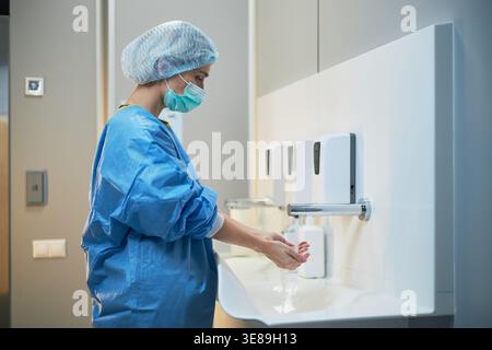 Junge Erwachsene kaukasische Chirurgin wascht vor dem Eingriff die Hände, unter Beachtung des Hygieneprotokolls und der Infektionskontrolle. Seitenansicht für Sicherheitsschulungen im Gesundheitswesen und medizinisches Marketing Stockfoto