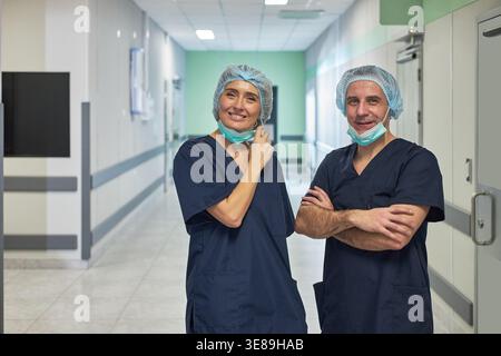 Porträt kaukasischer Chirurgen mittleren Alters, die im Krankenhauskorridor stehen, nach dem Eingriff lächeln und Masken senken. Nützlich für Personal im Gesundheitswesen, Teamarbeit, chirurgische Förderung Stockfoto