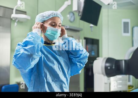 Junge Erwachsene Chirurgin, die Schutzmaske und Kappe im Operationssaal verstellt, die Vorbereitung auf den Eingriff und den sterilen Arbeitsablauf. Ideal für Personal im Gesundheitswesen, Chirurgie, Sicherheitsschulungen Stockfoto