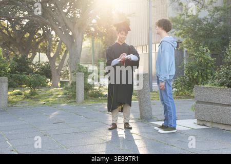 Verschiedene Männer stehen zur goldenen Stunde im Quad, Absolventen tragen Kleid und Mütze mit gerolltem Diplom. Innenhof, Campus, sonnenbeleuchtet, hintergrundbeleuchtet, im Freien, Feier, Freundschaft Stockfoto