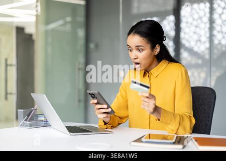Birassische Frau am Schreibtisch, die schockiert auf ihr Smartphone schaut, während sie eine Kreditkarte oder einen Laptop offen hält, auf unerwartete Gebühren für Online-Banking oder potenzielle Betrugsbenachrichtigungen reagiert Stockfoto