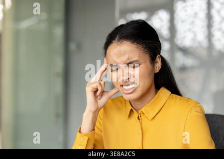 Die junge Frau hat schmerzhafte Kopfschmerzen, hält den Kopf und schließt die Augen, drückt Unbehagen, Stress und Leiden aus, während sie im Büro sitzt Stockfoto