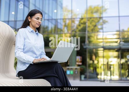 Junge indische Geschäftsfrau, die auf einer modernen Bank vor einem Glasgebäude sitzt, auf ihrem Laptop schreibt und sich auf Arbeit und Produktivität in einem professionellen städtischen Umfeld konzentriert Stockfoto