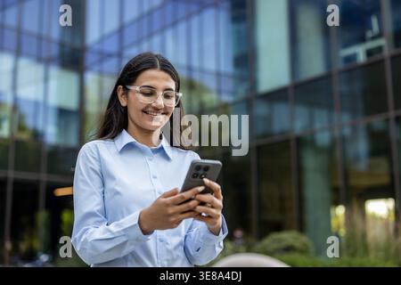Junge professionelle Frau lächelt fröhlich, während sie mit ihrem Smartphone interagiert, Nachrichten liest, surft oder an einer mobilen Anwendung in einer modernen städtischen Umgebung arbeitet Stockfoto