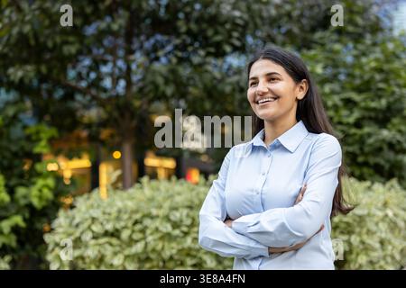 Indische Geschäftsfrau steht selbstbewusst mit gekreuzten Armen im Freien, lächelt und blickt weg und steht für Professionalität, Erfolg und positive Aussichten in einem Unternehmensumfeld Stockfoto