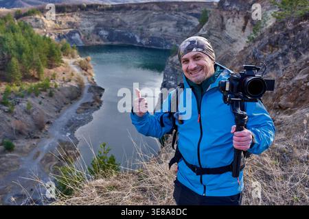 Ein lächelnder männlicher Videograf mittleren Alters, der die Daumen hoch hält, während er eine Kardankamera am Rande des Racos Steinbruchs hält, wobei der türkisfarbene See in der Ba sichtbar ist Stockfoto
