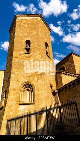 Flacher Blick auf einen historischen Steinkirchturm und eine Fassade hinter Eisenzaun unter blauem Himmel mit verstreuten Wolken, warmem Sonnenlicht und starken Schatten. Stockfoto