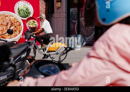 Taipeh, Taiwan - 28. April 2024: Maskierter Senior auf einem YouBike fährt den Bürgersteig, während ein Roller mit rosa Jacke parallel auf der Straße fährt. Stockfoto