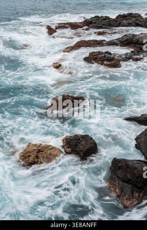 Dynamisches Ozeanwasser mit weißem Schaum wirbelt um zerklüftete, dunkle vulkanische Felsen entlang der Küste Stockfoto