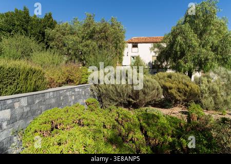 Eine lebendige Gartenlandschaft mit verschiedenen grünen Sträuchern und Bäumen umgibt ein klassisches weißes Gebäude mit gekacheltem Dach, das sich vor einem klaren blauen Sch befindet Stockfoto