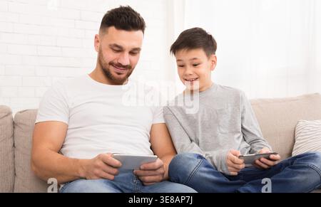 Vater und Sohn genießen die gemeinsame Zeit, während sie zu Hause Spiele auf ihren Geräten spielen Stockfoto
