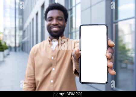 Lächelnder Mann, der das Smartphone vertikal mit leerem weißen Bildschirm für das Mockup hält, im Freien neben einem modernen Bürogebäude steht und mobile Apps, Werbung und digitale Displaykonzepte präsentiert Stockfoto