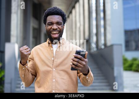 Glückliche Person, die auf einem Mobiltelefon gute Nachrichten erhält, Aufregung und Sieg ausdrückt, während sie vor einem modernen Gebäude steht und Erfolge und Triumphe feiert Stockfoto