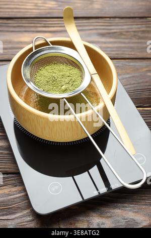 Sieb mit Matcha-Pulver und Schüssel auf Waage am Holztisch Stockfoto