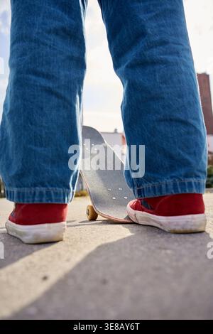 Niedriger Winkel von hinten auf eine Person in Jeans und roten Sneakers, die in der Nähe des Skateboards auf der Vorstadtstraße steht. Konzept des Skateboardens, des aktiven Lebensstils, des Trainings und des Outdoor-Hobbys. Stockfoto