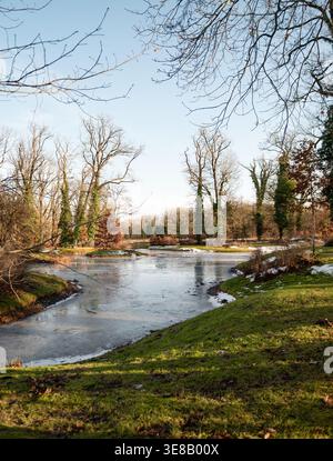 Gefrorener See und Winterlandschaft im Park. Malerischer Blick auf einen teilweise gefrorenen See, umgeben von Winterbäumen und grünem Gras. Der Winter wechselt in den Frühling Stockfoto