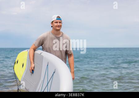 Der junge Mann steht mit seinem Stand Up Paddleboard am Wasser und genießt Outdoor-Fitness und Sommererholung im Meer. Stockfoto