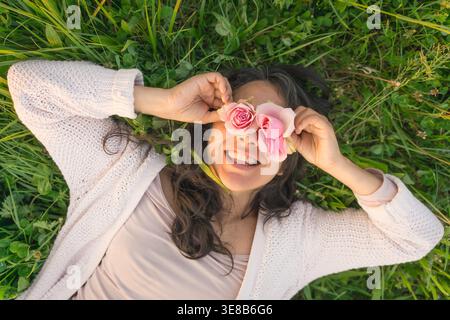 Junge Millennial Frau mit Blumen vor den Augen, die auf Gras auf der Wiese liegt, Rosen hält, hat Frühlingsstimmung, genießt angenehmen Geruch im Frühling Stockfoto