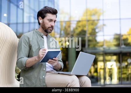 Mann mit schnurlosen Ohrstöpseln, die sich auf die Arbeit konzentrieren, in einem Notebook schreiben und ein Notebook verwenden, während er im Freien in einer städtischen Umgebung in der Nähe eines modernen Gebäudes sitzt und flexibles Arbeiten ermöglicht Stockfoto
