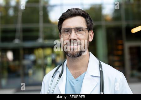 Bärtiger männlicher Arzt in weißem Mantel und Brille steht selbstbewusst vor einer modernen Klinik und lächelt in die Kamera, um professionelle, vertrauenswürdige Gesundheitsversorgung und patientenorientierte Pflege zu vermitteln Stockfoto