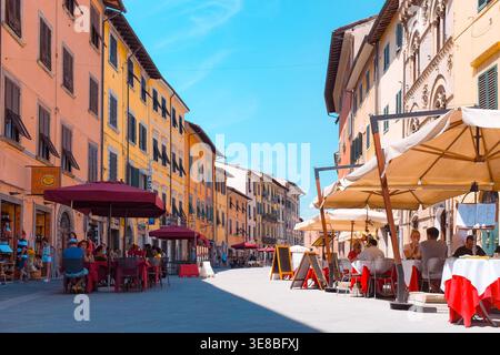 Pisa, Italien: Touristen und Einheimische essen im Freien in einer Reihe von Straßencafés mit weißen und roten Sonnenschirmen in einer sonnendurchfluteten historischen Straße. Stockfoto
