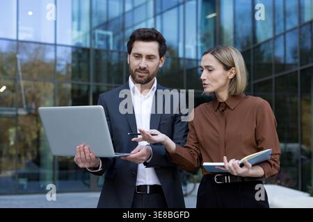 Geschäftskollegen arbeiten an einer Präsentation im Freien zusammen, prüfen Daten auf einem Laptop und teilen Ideen vor einem modernen Firmengebäude, wobei Teamarbeit und Strategie zum Ausdruck kommen Stockfoto