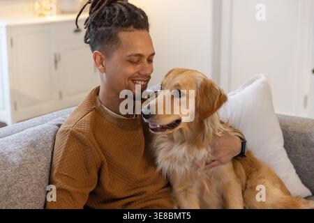 Mann mittleren Alters, der auf einem grauen Sofa sitzt und einen goldenen Retriever hält, mit braunem gestepptem Sweatshirt Stockfoto