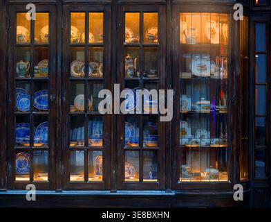 Ein wunderschön gefertigter antiker Holzschrank mit Glastüren, gefüllt mit einer Auswahl an bunten Tellern, Geschirr und Teekannen auf mehreren Regalen, Stockfoto