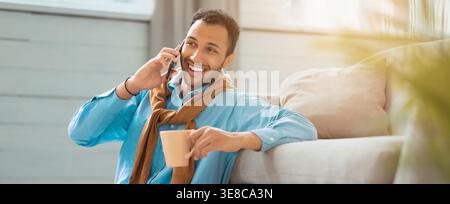 Mann spricht am Telefon, während er auf einer Couch sitzt und einen Becher in einem hellen Raum hält Stockfoto