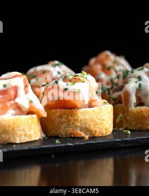 Zarte Lachsscheiben ruhen auf geröstetem Brot. Cremige Soße tränkt kunstvoll über jedes Stück. Frische Kräuter garnieren die Vorspeise mit leuchtendem Grün. Stockfoto