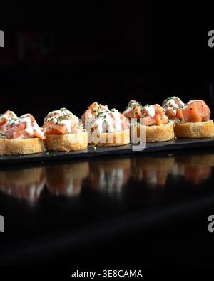 Salzige Lachsscheiben stehen auf geröstetem Brot. Frische Kräuter und cremige Soße sorgen für einen optischen Reiz. Dunkle Platte steht im Kontrast zu hellen, einladenden Farben. Abendessen- Stockfoto