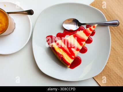 Ein Stück Käsekuchen mit frischer Erdbeermarmelade, serviert auf einem grauen Teller neben einem Glas Kaffee in der Cafeteria Stockfoto