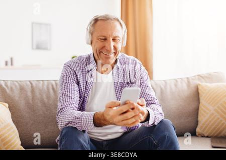 Ein älterer Mann nutzt das Smartphone gern, während er Musik im gemütlichen Wohnzimmer hört Stockfoto