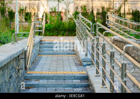 Eine Treppe mit grauen Steintreppen und Metallgeländern auf beiden Seiten für Zugänglichkeit, die zu einer höheren Ebene mit Grün und einem Gebäude im führt Stockfoto