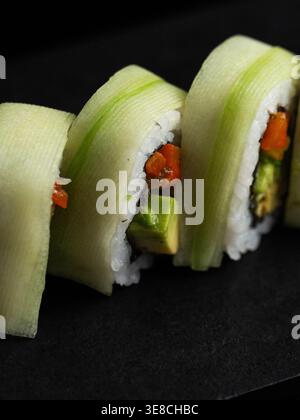 Sushi-Brötchen in Gurkenscheiben gewickelt. Leuchtend orange und grüne Füllungen sehen durch. Weißer Reis steht im Kontrast zu dunkler Oberfläche. Nahaufnahme zeigt Text Stockfoto