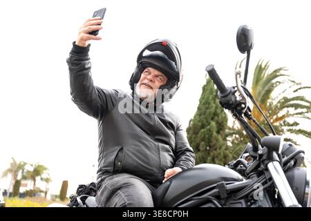 Erfahrene Motorradfahrer machen ein Selfie mit Smartphone, genießen Freizeitreisen auf einem Motorrad, tragen Helm und Lederjacke im Freien Stockfoto