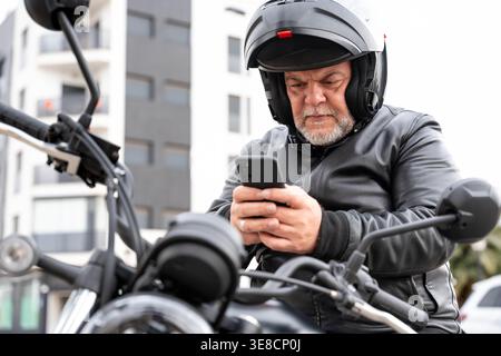 Ein erfahrener Biker, der auf seinem Motorrad saß, eine Lederjacke und einen Helm trug, konzentrierte sich auf sein Mobiltelefon in einer urbanen Umgebung Stockfoto