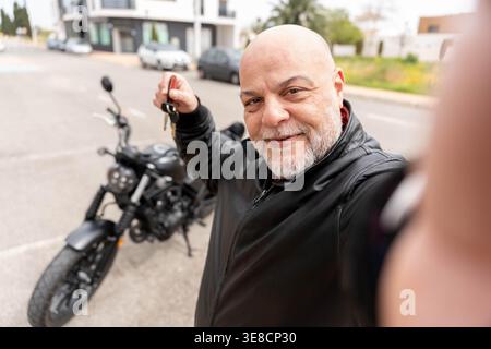 Bärtiger Glatzkopf, der die Schlüssel zu seinem neuen dunklen Motorrad hält und sich glücklich fühlt Stockfoto