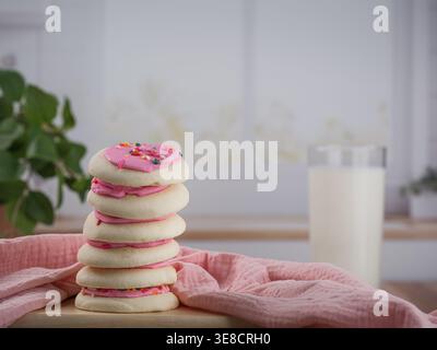 Stapel rosafarbener, mattierter Frühlings-Osterzucker-Kekse mit hohem Glas kalter Milch und Stoffserviette aus Musselin-Stoff auf Holztisch mit Pflanze Stockfoto