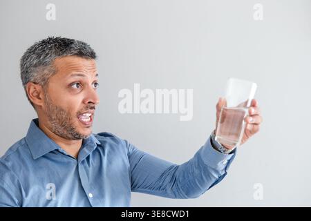Schockierter Mann mittleren Alters in einem blauen Hemd, der ungläubig auf ein klares Glas Wasser blickt, das er vor weißem Hintergrund hält Stockfoto