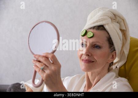 Eine Frau mit einem Handtuch auf dem Kopf entspannt sich in einer Spa-ähnlichen Umgebung. Sie hält einen kleinen Spiegel und blickt sich mit Gurkenscheiben auf der Stirn an. Stockfoto