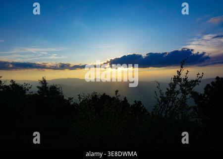 Sonnenuntergang hinter einer Bergkette mit goldenem Licht, das durch Wolken über ein üppiges Tal strömt. Stockfoto