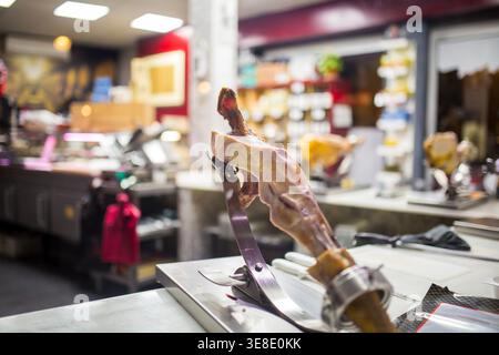 Ein köstliches serrano-Bein mit geräucherter spanischer Marmelade wird auf einem traditionellen Schnitzerstand in einem belebten Gourmet-Geschäft montiert, bereit für professionelles Schneiden und en Stockfoto