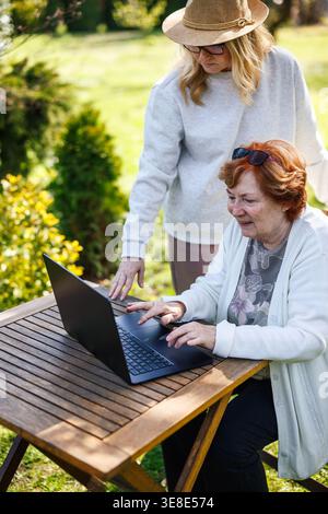 Ältere Mutter lernt digitale Fähigkeiten und moderne Technologie von ihrer erwachsenen Tochter. Ältere Frau, die im Freien einen Laptop benutzt Stockfoto