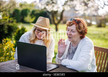 Ältere Frau und Erwachsene Tochter haben Videoanrufe auf Laptop im Frühjahrsgarten. Zwei Frauen lachen und kommunizieren im Freien über das Internet Stockfoto