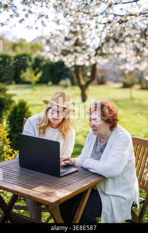Erwachsene Tochter, die älteren Müttern die Benutzung eines Laptops beibringt. Frauen sitzen am Tisch unter blühenden Kirschbäumen im Frühlingsgarten Stockfoto